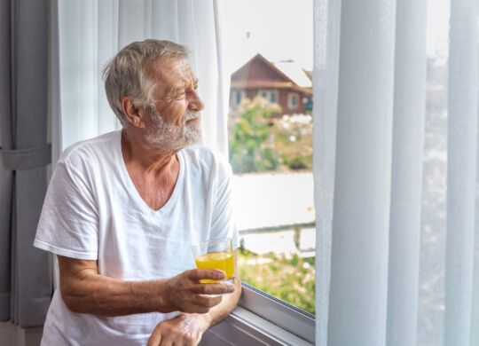 Senior elderly man standing and looking out of window in bedroom after waking up in morning with juice