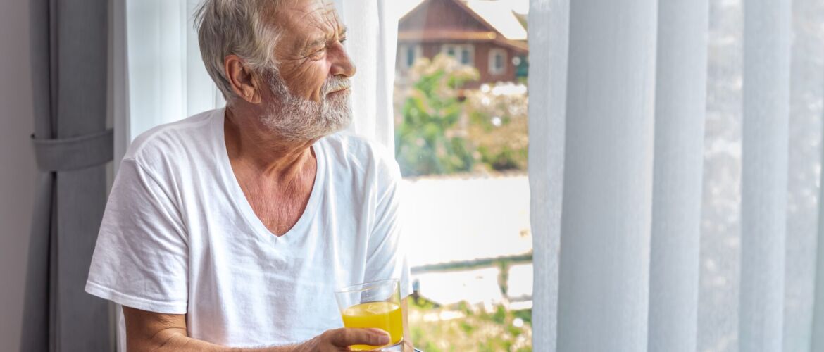 Senior elderly man standing and looking out of window in bedroom after waking up in morning with juice